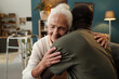 © pressmaster - Senior Caucasian woman hugging Black man in living room, both showing affection and warmth, elderly woman smiling with eyes closed, walker visible in background