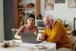 © pressmaster - Young adult Hispanic woman assisting senior Caucasian man using laptop at kitchen table, both focusing on screen while discussing documents and working together on paperwork