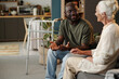 © pressmaster - Black man sitting on sofa talking with senior Caucasian woman, both engaged in conversation in home setting, walker visible in background indicating mobility assistance