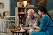 © pressmaster - Senior Caucasian man sitting while young adult Latin woman in medical scrubs measuring blood pressure using digital monitor, walker and plants visible in background