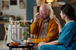 © pressmaster - Senior Caucasian man drinking water while sitting beside young adult Latin woman in medical scrubs, caregiver gently touching his shoulder, walker and breakfast tray visible nearby