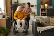 © pressmaster - Senior Caucasian man with disability sitting in wheelchair talking to young adult Latin woman standing behind him in living room, surrounded by houseplants and cozy decor