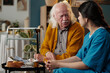 © pressmaster - Senior Caucasian man sitting and talking with young adult Latin woman in medical scrubs, holding cane and surrounded by medication and food on table in healthcare setting