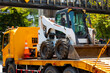 Skid steer loader on flatbed truck in urban with traffic and greenery