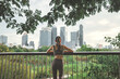 Rear view of Asian woman in sportswear taking a break after running in Bangkok park at early morning, looking at city skyline in distance, symbol of healthy urban lifestyle and wellness