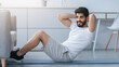 © Prostock-studio - Training in covid-19 quarantine, sports workout, gym at home and healthy lifestyle. Glad arabian bearded young male doing abs exercises in living room interior, on mat with dumbbells on floor
