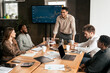 © Prostock-studio - Corporate Meeting Concept. Smiling Male Entrepreneur Giving Speech During Briefing With Coworkers Employees In Modern Office, Standing At Table In Board Room, Discussing Strategy And Management