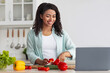 © Prostock-studio - Online lesson, food class remotely, new normal and modern cooking. Smiling millennial african american female cuts pepper and looks at laptop in kitchen interior with bright vegetables on table