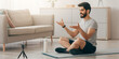 © Prostock-studio - Sports blogger, online trainer and remote training, yoga practice. Happy arabian millennial male bearded coach sits on mat and talks with followers, looks at webcam on phone in living room interior