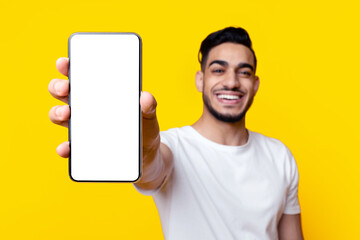 Happy handsome middle-eastern young man showing modern smartphone with empty screen, recommending nice mobile application or showing amazing offer over yellow studio background, mockup