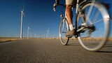 Medium shot of a cyclist riding past wind turbines clear focus on the bike and rider with blurred white blades in motion symbolizing clean transportation and wind energy.