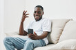 © SHOTPRIME STUDIO - Happy African man enjoying video call on smartphone, sitting on a comfortable couch in a bright, modern living room, in casual white t shirt and blue jeans