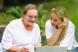 © Iryna - Enjoying a sunny afternoon, two women share moments of joy while exploring a laptop outdoors in a serene garden setting