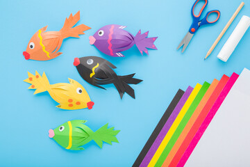 Wall Mural - Fishes, scissors, glue stick, pencil and colorful application paper on light blue table background. Pastel color. Closeup. Child created shapes. Top down view.