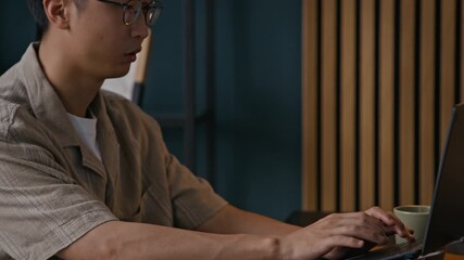 Wall Mural - Handheld shot of male Asian freelancer getting tired of working on laptop while programming at table in cozy home office