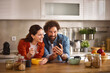 © Stockphotodirectors - A couple sits at a wooden table in their home office, sharing a bowl of cereal. They laugh as they look at a smartphone, creating a warm and joyful atmosphere.