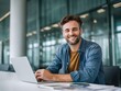 © Yuliia Malovana - Smiling businessman in casual clothes working on a laptop at desk with modern office background and looking at the camera. Happy satisfied male employee typing and doing his project on a computer.