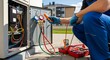 © NanzZ - Technician wearing blue overalls and gloves is servicing an air conditioning unit, using gauges and a multimeter to check the system.
