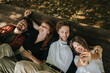 © qunica.com - A group of four friends share a happy moment outdoors, lying together on a wooden bench under shaded trees, enjoying nature and camaraderie, with one capturing the moment in a selfie.