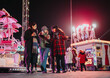 © qunica.com - Three friends gather at night in a lively holiday fairground setting, enjoying warm beverages and festive cheer among bright lights and rides, embodying the warm spirit of winter festivities.
