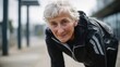 © Tina - Senior Caucasian woman with short gray hair, wearing a black athletic jacket, smiling while leaning forward outdoors. The scene conveys fitness and active lifestyle.