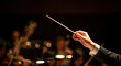 © Len_mik - Conductor's hands leading orchestra with baton in lighted concert hall. Hands conducting orchestra include skillful gestures, focused musicians in background creating harmonious sounds.