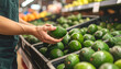 © Athiphat - Shopper selecting fresh avocado in grocery store, vibrant green fruit displayed in produce section, healthy food choice, market atmosphere, natural lighting, selective focus