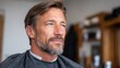 © SerPak - A man with a well-groomed beard sits in a barber shop, looking thoughtful. The setting is relaxed and professional, ideal for grooming services.