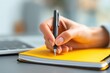 © Alohanongza - A hand holds a pen and writes in a yellow notebook, with a laptop in the background on a desk