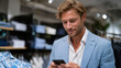 © Milos - A charming man checks his phone while surrounded by fashionable clothing, illustrating the blend of modern technology and personal style in a contemporary shopping experience.