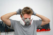 © Mihail - male person in a gray shirt puts on a mask in the paint shop, emphasizing safety precautions while preparing for painting tasks.