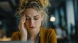 © Aliaksandr - A young Caucasian woman with curly blonde hair looks stressed while sitting at a desk in an office environment. She has her hand on her forehead, indicating frustration.