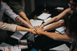 © qunica.com - A group of coworkers join hands above a workspace, symbolizing unity and collaboration. The setting indicates a collaborative effort during a team meeting or brainstorming session.