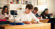 © peopleimages.com - Boy, student and writing with books in classroom for higher education, learning or development. Male person, teenager and studying with textbooks in class for knowledge, assessment or test in school