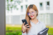 © maeching - Asian female student in warm clothes using phone to chat with friends and ready for video call while in university.