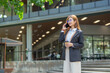 © maeching - Smiling Asian businesswoman in suit holding coffee cup and documents walking in front of modern business building