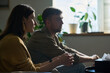 © pressmaster - Caucasian young adult woman and Caucasian young adult man sitting side by side, focusing intently on something out of frame, hands clasped, natural light coming through window