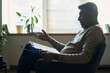 © pressmaster - Middle aged Indian man sitting in chair holding clipboard and eyeglasses, gesturing with hand while engaging in conversation, indoor office setting with window and plants visible