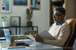 © pressmaster - Middle aged Indian man sitting at desk reviewing documents while working on laptop in modern office, wearing eyeglasses and concentrating on paperwork, indoor business setting