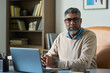 © pressmaster - Portrait of middle aged South Asian man with gray hair and beard sitting at desk using laptop, wearing eyeglasses, looking into camera, bookshelf in background