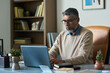 © pressmaster - Middle aged Indian man working on laptop at desk in modern office, wearing glasses and beard, focusing on screen, surrounded by books and plants, appearing concentrated