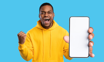 Emotional african american guy gamer showing smartphone with blank screen and celebrating success, trading online, using mobile application, blue studio background, panorama, copy space, mockup