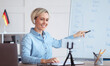 © Prostock-studio - Online languages school. Millennial female tutor giving German lesson on internet, using smartphone, pointing at blackboard, explaining grammar to remote students. Education during covid-19 epidemic