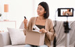 © Prostock-studio - Product Review. Satisfied asian young woman vlogger holding foundation and brush, unpacking cardboard beauty box with cosmetics products, recording video for her social media blog on camera at home