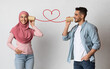 © Prostock-studio - Romantic arab guy sending love message to muslim girlfriend through paper cup phone connected by heart shaped string, standing together over grey studio background, collage for Valentine's day