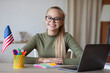 © Prostock-studio - Cute teen girl with USA flag using laptop, studying american english online from home, copy space. Smiling female teenager having educational online course, foreign languages learning