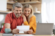 © Prostock-studio - Happy middle aged spouses reading letter from bank, sitting at table in front of laptop, drinking coffee, checking correspondence together, kitchen interior, copy space. Mortgage, loan concept