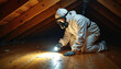 © Vadym - Man in protective suit with mask, goggles inspects attic floor using flashlight. Cautious home maintenance for safety, pest control, or renovation. Worker kneels on wooden planks checking for issues.
