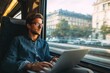 © ant - Young professional sitting in a moving train or bus, working on laptop, city view through window