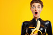 © alexkoral - surprised sexy young woman with mouth open in a black latex suit holds a big banana on a yellow isolated background. Symbol of oral sex with a male large penis